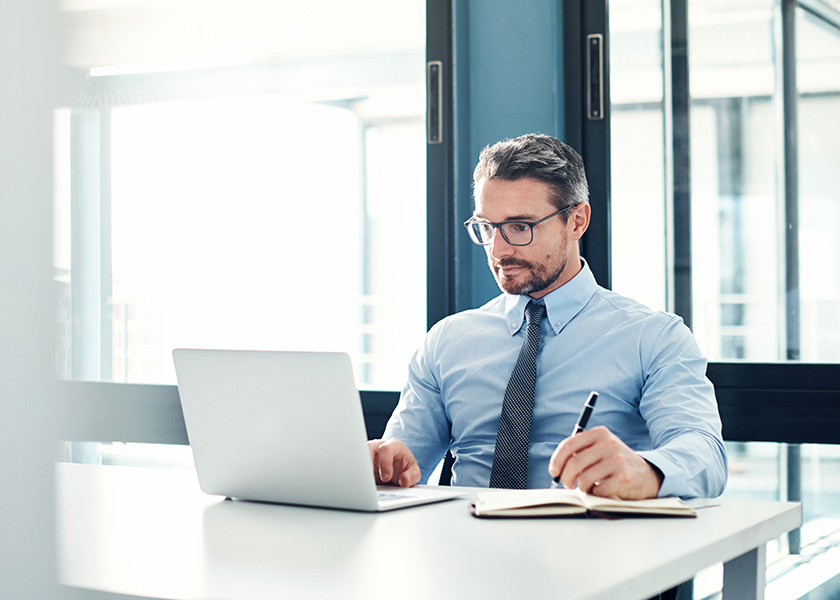 A man in a tie sits at a conference table working on a laptop, with a pen and paper nearby. Business owner planning to sell.